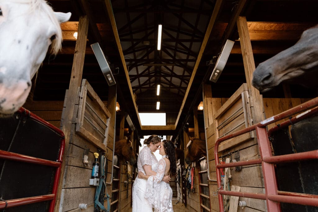Two brides embracing in a barn with horses on each side.