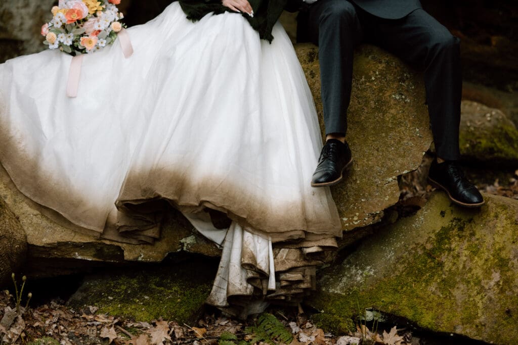 Close up of bride's dress and groom's pants and shoes while they are sitting on a rock. 