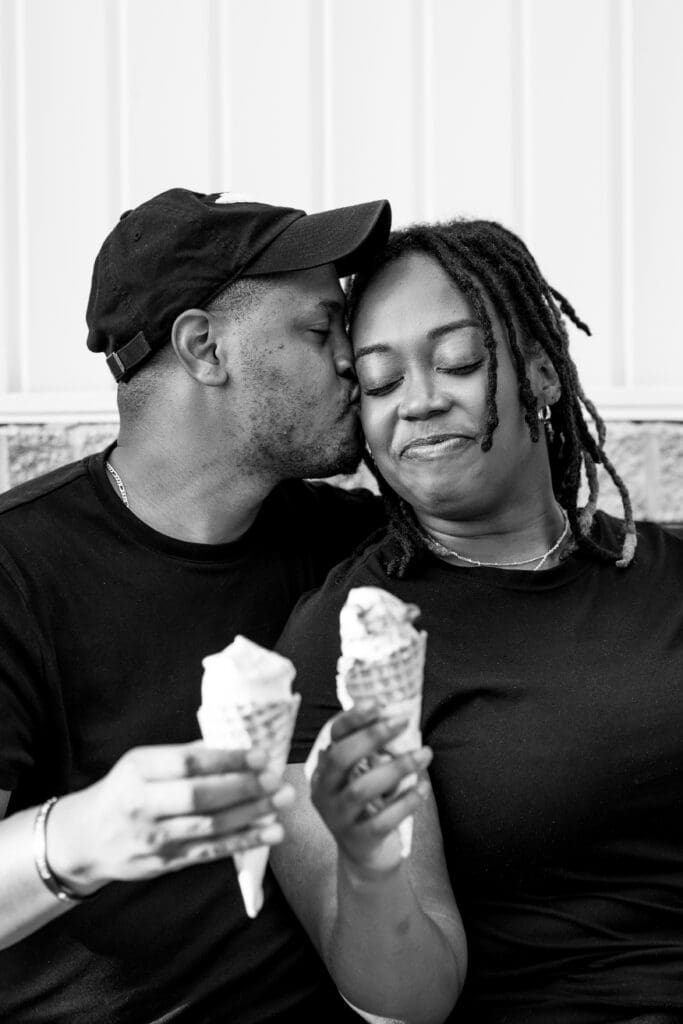 Man kissing girlfriend on her cheek while they hold ice cream.