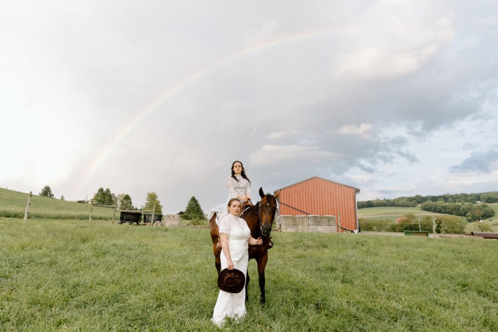 Two brides, one on a horse, one in front of the horse. A rainbow is in the sky behind them.