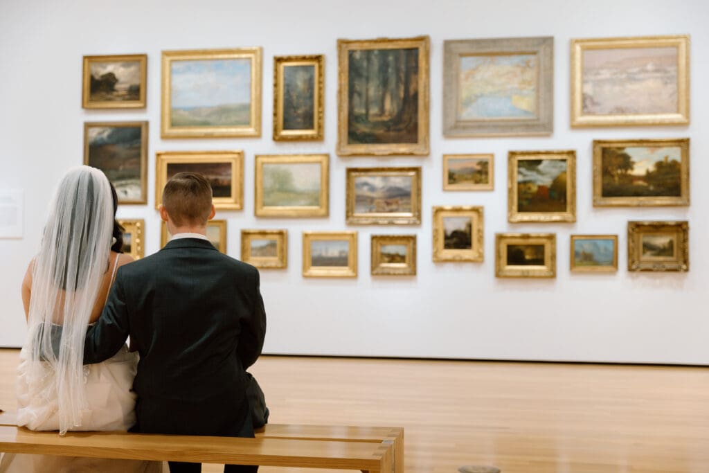 Bride and groom sitting on art gallery bench and looking at gallery display.