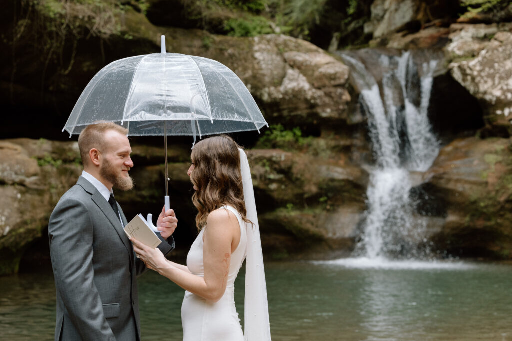 Elopement couple reading vows under a clear umbrella in front of upper falls at Old Man's Cave.