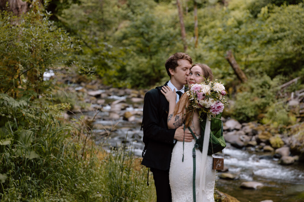 Couple embracing while the bride looks back. A river is flowing behind them.