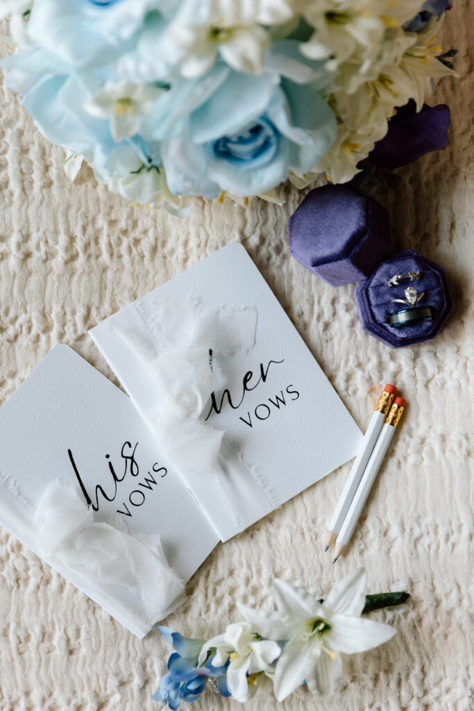 Details photo with vow books, pencils, flowers, and rings.