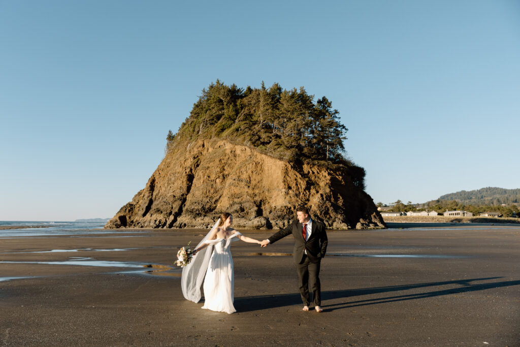 Couple holding hands and walking with a sea stack behind them on the Oregon Coast at sunset.