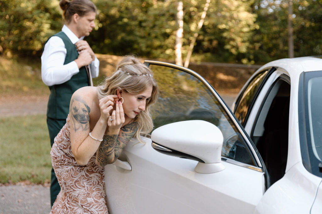 Bride adjusting earring in car mirror while groom is getting dressed behind her.
