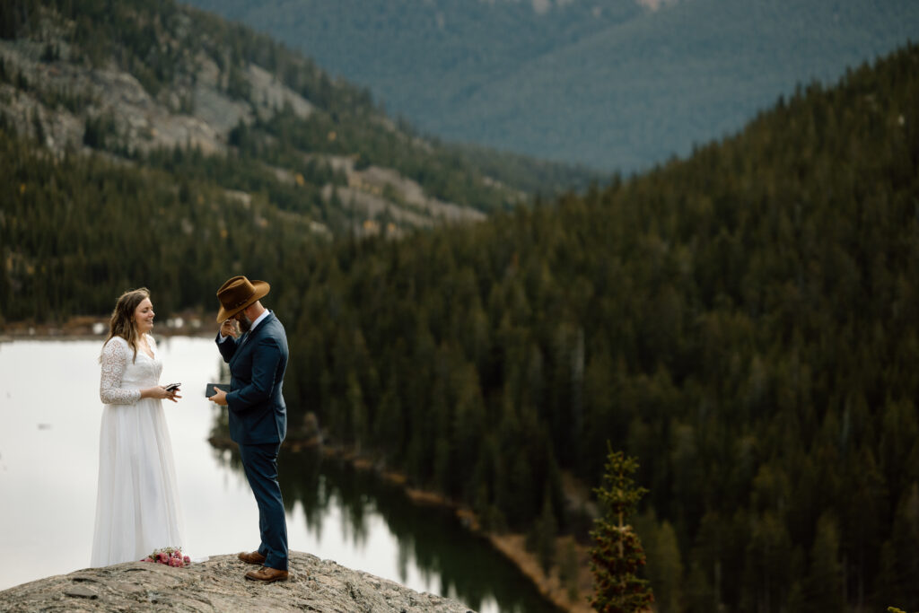 Couple standing on a rocky outcrop in front of an alpine lake surrounded by green trees. 
