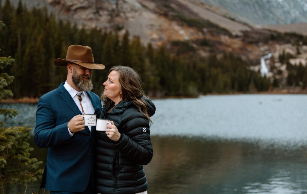 Couple clinking coffee mugs in front of an alpine lake.