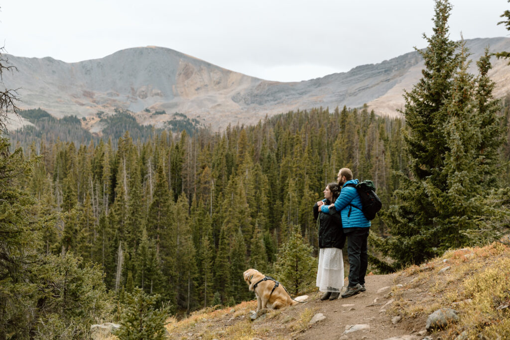 Elopement couple embracing while their dog sits in front of them. Mountains and trees are in the background.