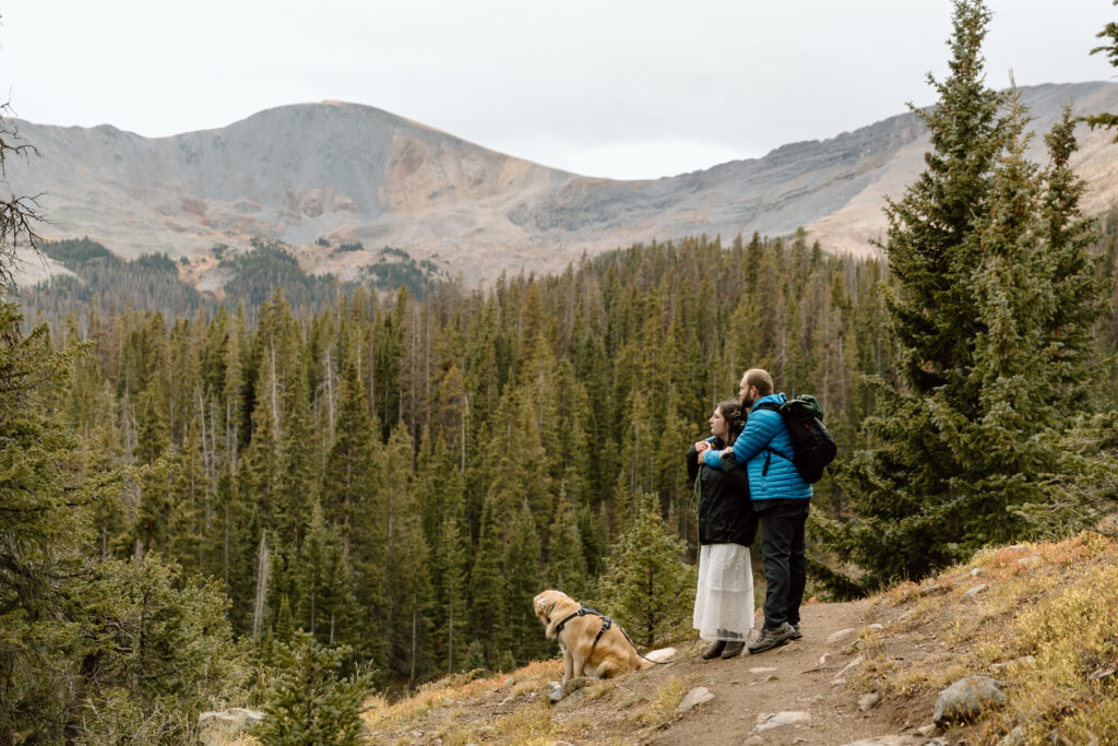 Couple embracing and looking out on the mountains. Their dog is sitting in front of them.