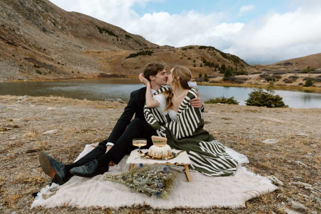 Couple kissing while seated on a picnic blanket and sharing a cake.