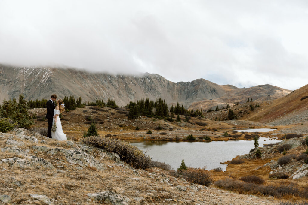 Couple embracing on a hill in front of an alpine lake with mountains in the background.