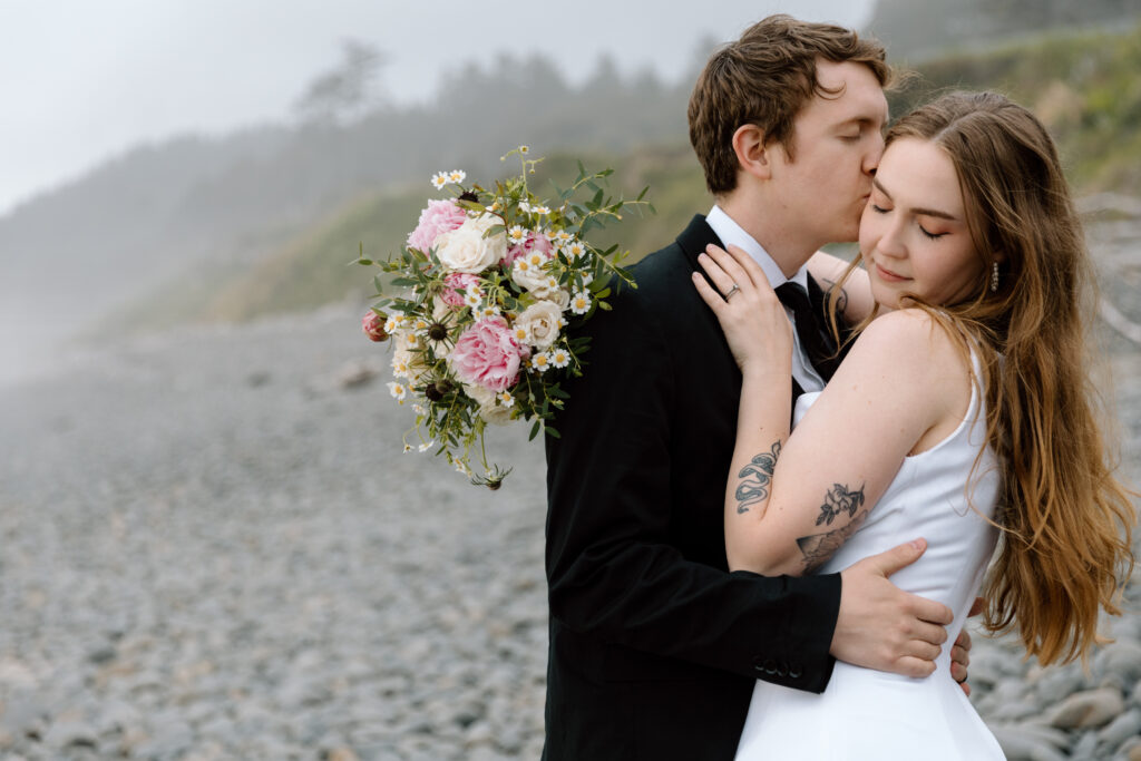 Couple embracing while groom kisses bride on cheek. They are on a rocky Oregon Coast beach.