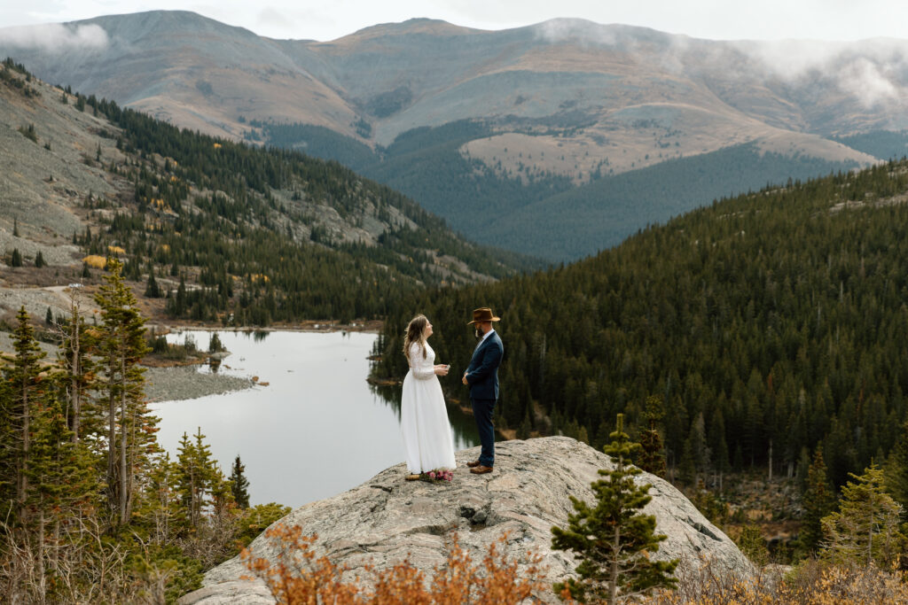 Couple standing across from each other on a rocky outcropping with mountains behind them.